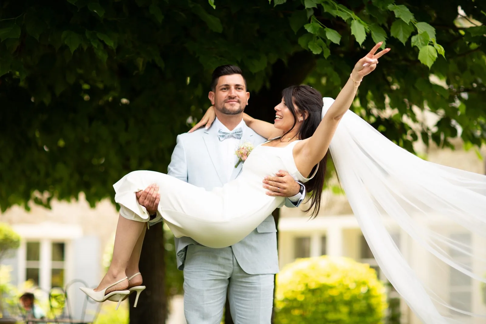 Le marié porte la mariée dans ses bras sous les arbres, son voile au vent