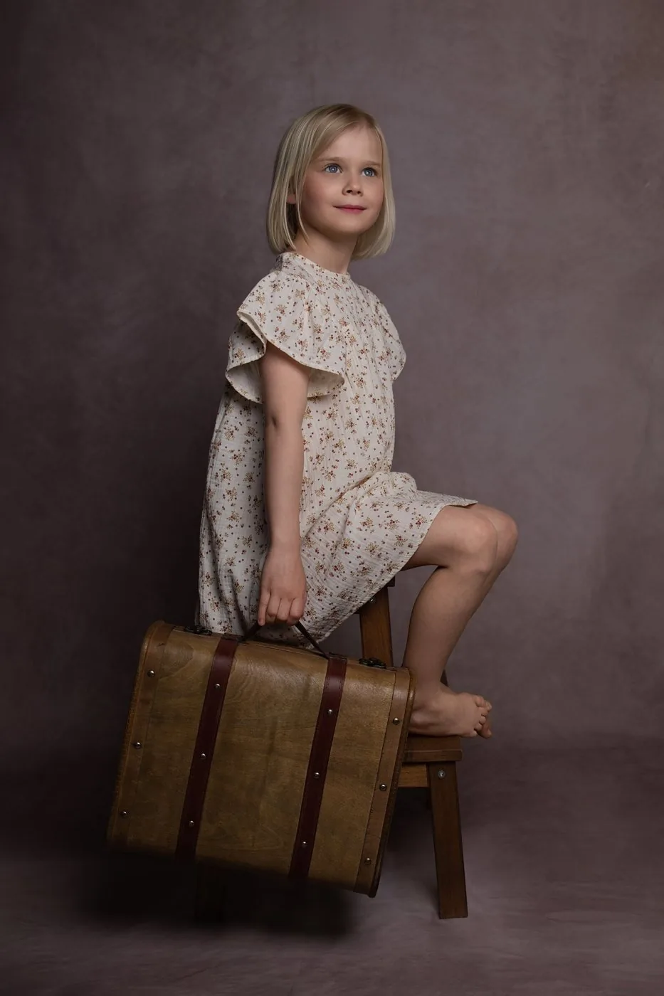 Assise sur un tabouret, la fillette tient une valise et lève les yeux en souriant