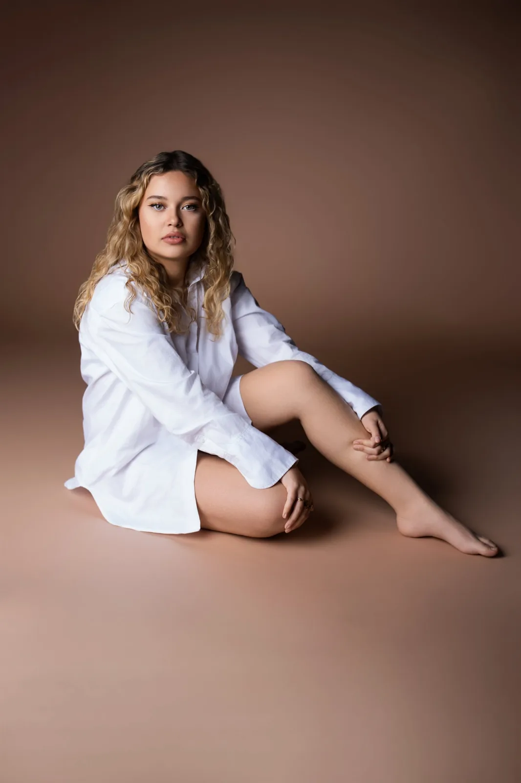 Jeune femme aux cheveux bouclés assise au sol en chemise blanche, fond terracotta